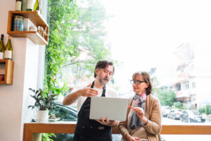 Cafe owner consults with another small business owner using laptop in a bright cafe setting. Their collaboration represents the opportunity to boost your sales by partnering with other businesses.