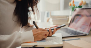 A person in a white sweater creates a social media calendar, writing notes in a notebook while holding a smartphone, with a laptop nearby.
