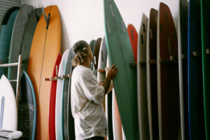 A person with long hair and a cap inspects a green surfboard among various other colorful surfboards on racks. Get insights on managing a family business transfer from the owners of several successful surf shops.