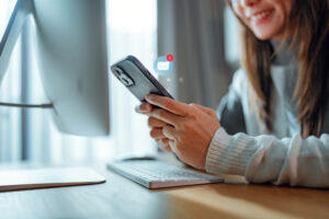 A woman smiling while holding a smartphone with a notification icon, sitting at a desk with a computer monitor and keyboard, representing email marketing for small businesses 2025.