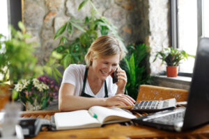 Smiling small business owner managing finances with a calculator and notebook while talking on the phone, surrounded by plants, symbolizing effective cash flow management for small businesses in a relaxed work environment.