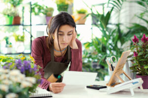 A small business owner, seated at a white desk in a plant shop, reviews paperwork with a thoughtful expression. The desk is surrounded by vibrant potted plants, a calculator, and a tablet stand, symbolizing the use of fintech solutions for small businesses to manage finances and grow operations.