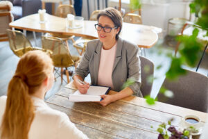 Two women engaged in a professional one-on-one meeting at a wooden table in a relaxed cafe setting, symbolizing effective employee reviews for small businesses. One woman smiles while taking notes on a clipboard, emphasizing open communication and constructive feedback.