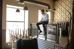 A modern café with stacked chairs on tables, pendant lights, and a man sitting on a raised platform using his smartphone. The scene reflects a small business managing fixed costs by optimizing space and resources during non-operational hours.