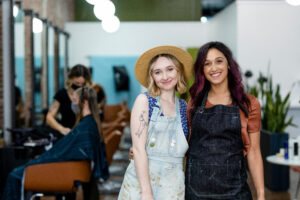Two smiling Gen Z employees in a modern hair salon, standing together with a friendly pose, showcasing teamwork and a welcoming workplace environment.
