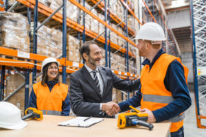 Three individuals in a warehouse setting engage in a positive business interaction. Two workers wearing white hard hats and orange safety vests stand beside a man in a suit, shaking hands and smiling. A table in the foreground holds a white hard hat, scanning devices, and a clipboard, emphasizing workplace operations. The industrial background features tall shelves stacked with boxes and pallets. This scene illustrates collaboration and success, highlighting the importance of employee incentive programs for small businesses in fostering teamwork and productivity.