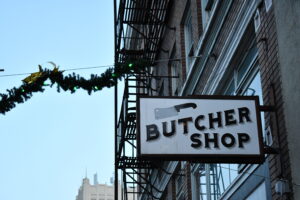 A festive street scene featuring a 'BUTCHER SHOP' sign hanging from a brick building, with a fire escape and holiday garland in the background. This image highlights the importance of exterior signage for small businesses in attracting customers and enhancing visibility.