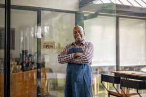 A small business owner in a plaid shirt and blue apron stands confidently with arms crossed in front of their cafe, symbolizing success and preparation for the future, aligning with the concept of how to retire as a small business owner.