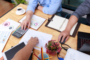 Hands of three business partners working together on cost analysis exercises, using charts, a calculator, and a laptop.