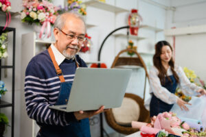 A senior man stands in a flower shop, using a laptop to apply for small business grants, while a young woman carefully wraps flowers in the background.