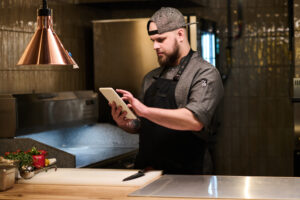 A male chef and small business owner wearing a gray apron checks his tablet after improving his pizzeria's local search results.