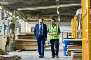 A young workman wearing a safety vest gives a tour of a factory to a suited businessman considering the pros and cons of local vs. global suppliers for his small business.