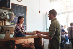 A woman in an apron hands a coffee cup to a man at a café counter. Research shows that nearly 80% of consumers have a small business blind spot, greatly underestimating the number of small businesses in operation.