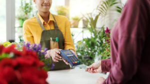 A shop attendant in a yellow shirt and green apron holds a card reader as a customer in a maroon shirt hands over a credit card in a vibrant plant shop. The image highlights a retail transaction, symbolizing the impact of credit card swipe fee reform on small businesses.