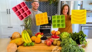 Ross and Tess Nelson, founders of Smootheeze, smiling in a modern kitchen while holding colorful silicone molds, surrounded by an array of fresh fruits and vegetables.