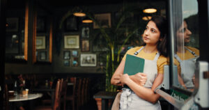 A waitress gazes thoughtfully out of a window, symbolizing the emotional and operational challenges businesses face due to the impact of tariffs.