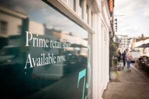 A storefront window with bold white text reading "Prime retail space" and "Available now," set against a dark reflective background. The scene includes a busy commercial street with pedestrians and shop facades, emphasizing a prime location. Perfect for illustrating lease negotiation tips in a bustling retail environment.