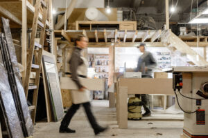 A busy woodworking workshop representing small business sectors hit hardest by tariffs. Two workers in motion, one carrying a wooden panel and another operating machinery, surrounded by tools, raw materials, and industrial equipment, highlighting the challenges faced by small businesses in affected industries.