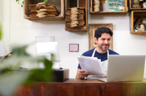 A smiling man, likely a small business owner, stands behind a wooden counter in his shop. He wears a dark blue t-shirt and a white apron, holding a stack of papers in one hand while interacting with a sleek silver laptop. A modern white point-of-sale system is visible on the counter. The background features rustic wooden shelves filled with neatly organized items like paper bags, twine, and decorative trinkets. The scene conveys a professional and welcoming atmosphere, representing the dedication and daily operations of small business owners. This image aligns with the theme of the Square small business grant, showcasing the hardworking individuals it aims to support.