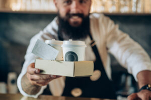 A barista in a coffee shop offers a cardboard tray with two takeaway coffee cups and a loyalty card featuring circular stamps, promoting small business loyalty programs.