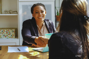 Two professional women shaking hands across a wooden table in a well-lit office setting, symbolizing a successful agreement or partnership. The scene conveys trust and collaboration, aligning with the concept of the best background check services for small businesses.