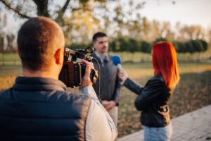 A cameraman records an interview between a journalist and a small business owner in a park. When considering how to get press coverage for your small business, reach out to blogs, websites, newsletters, podcasts, TV stations, and social media influencers.