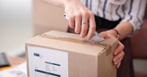 A close-up of hands opening a brown cardboard box with a utility knife, cutting through clear packing tape. The box features a delivery note label with order details. The person wears a striped shirt, a watch, and a ring. The image illustrates the end of the de minimis exemption in the United States and the resulting impact on small businesses.