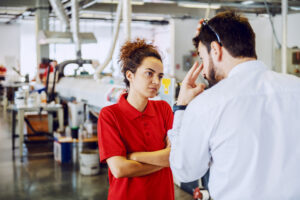 A woman in a red polo shirt and a man in a white collared shirt engage in a serious discussion in an industrial workshop setting. Their focused interaction highlights the need for clear workplace policies, such as an employee handbook for small business environments.