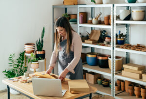 A woman, representing a small business owner, is seen in a bright, organized craft studio, carefully packaging a product at a wooden table. Surrounding her are potted plants and shelves filled with handcrafted pottery and supplies. This scene highlights the dedication and craftsmanship of small businesses pursuing B Corp certification for small business growth and impact.