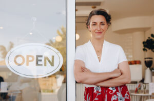 A small business owner stands confidently in the open doorway of her shop. She wears a white top and a red floral skirt, with an 'OPEN' sign hanging on the glass door beside her. The inviting scene reflects the theme 'how to market your woman-owned business,' emphasizing warmth, professionalism, and approachability.