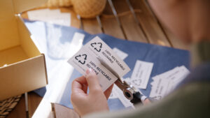 Close-up of hands cutting eco-friendly labels on a blue surface, featuring messages like '100% Recyclable' and 'Reusable,' emphasizing sustainability for small businesses. Scattered labels and a cardboard box in the background highlight a focus on sustainable packaging practices.
