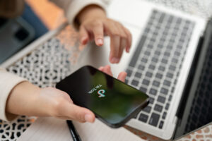 A close-up view of hands interacting with a smartphone displaying the TikTok app, positioned on a patterned surface alongside an open laptop. The image highlights the use of technology and social media platforms for small business operations, emphasizing engagement with digital tools like TikTok for marketing and outreach.