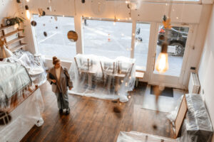 Overhead view of a commercial space undergoing renovation, with furniture covered in plastic sheeting and a woman holding a laptop standing on a wooden floor. The scene reflects preparation or transition, symbolizing investment and change, aligning with capital spending trends.