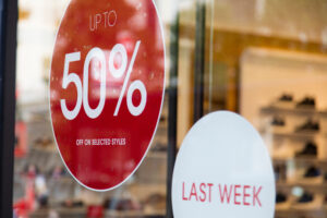 Storefront window with sale signs promoting holiday discounts for small businesses. A large red circular sign displays 'UP TO 50% OFF ON SELECTED STYLES,' while a smaller white oval sign reads 'LAST WEEK,' creating urgency for shoppers.