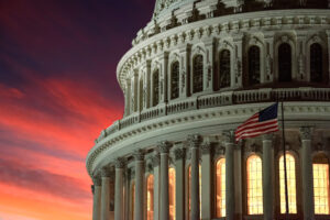 The U.S. Capitol building illuminated at dusk with a dramatic red and orange sunset sky in the background. An American flag waves prominently in the foreground. Here in Washington, D.C., Congress may be presented with an opportunity to advance small business healthcare reform.