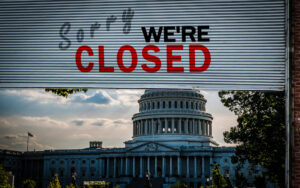 A metallic security shutter with the text 'Sorry WE'RE CLOSED' in bold black and red letters, similar to phrasing used by small businesses. Through the partially open shutter, the U.S. Capitol Building is visible under a cloudy sky, symbolizing the October 2025 government shutdown.