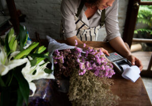 A florist or small business owner wearing a striped apron works at a wooden counter surrounded by fresh white lilies and dried flowers. They use a calculator with a receipt printer, symbolizing financial management and strategies for how to handle rising costs.