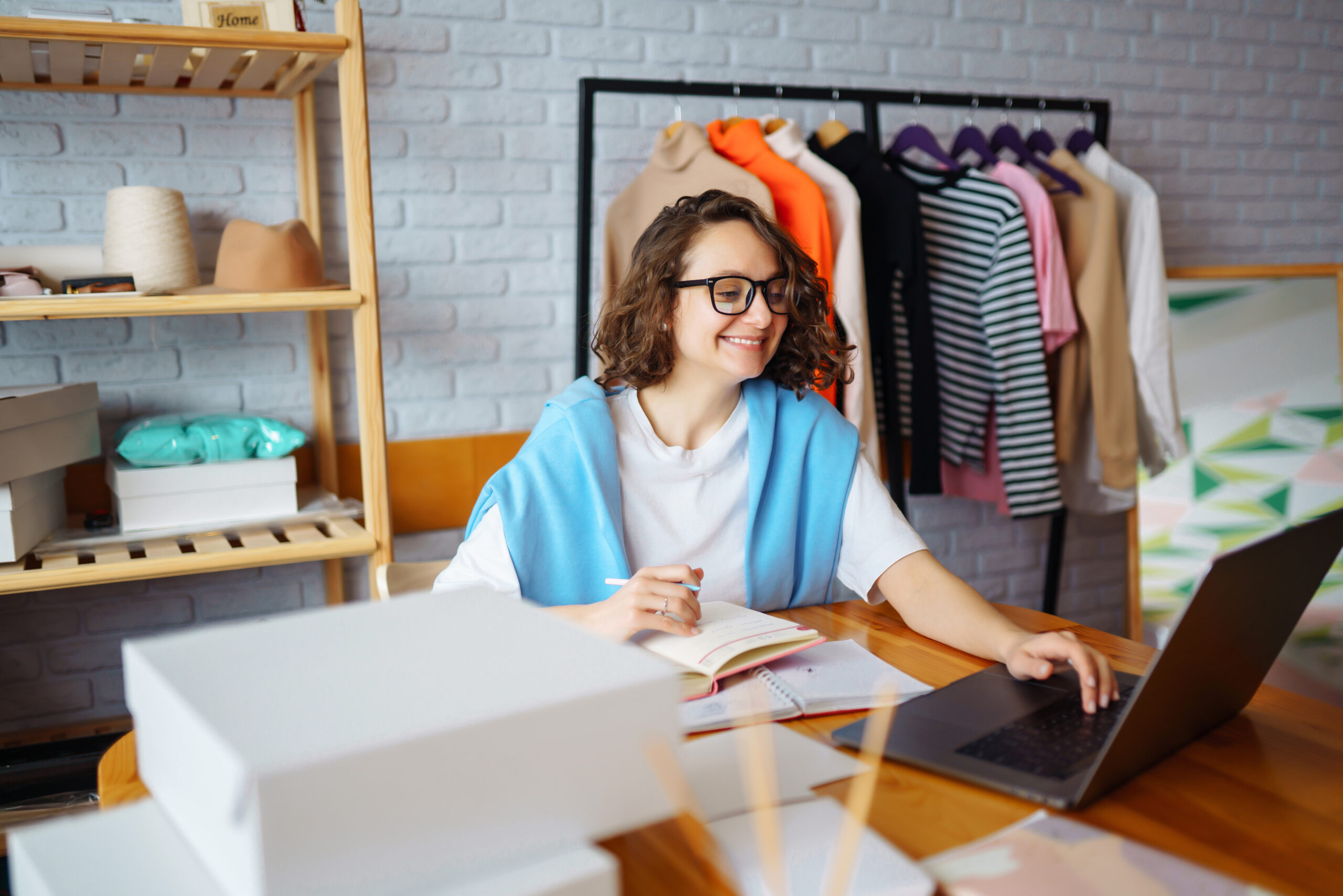 A smiling small business owner working at her desk in a bright, organized home office. She uses a laptop and notebook, surrounded by packaging boxes and a clothing rack, symbolizing affordable email marketing for small businesses in the fashion or e-commerce industry.