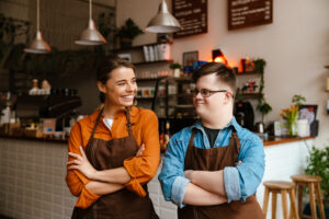 Two smiling baristas, a woman and a man with Down syndrome, stand side-by-side in a cozy coffee shop, both wearing aprons with arms crossed. The image highlights an inclusive workplace, showcasing the benefits of hiring people with disabilities in small businesses.