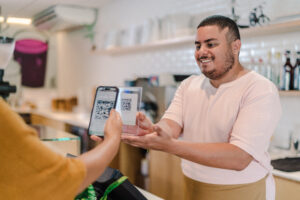 Customer scanning a QR code on a transparent display held by a smiling barista in a cafe, illustrating a modern transaction. This image highlights the importance of avoiding common QR code mistakes in small business operations.