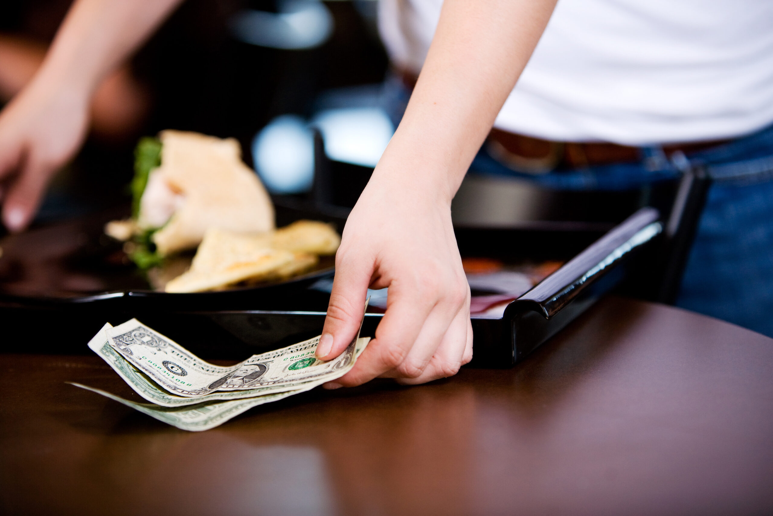 A close-up of a person's hand reaching for a small stack of dollar bills on a wooden table next to a black tray with food, suggesting a casual dining setting. The image represents tipping practices and financial transactions, aligning with the "tips deduction business savings" topic. The blurred background hints at a restaurant environment. A close-up of a person's hand reaching for a small stack of dollar bills on a wooden table next to a black tray with food, suggesting a casual dining setting. The image represents tipping practices and financial transactions, aligning with the "tips deduction business savings" topic. The blurred background hints at a restaurant environment.