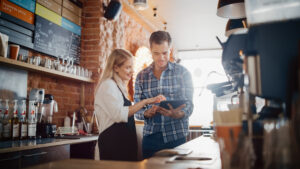 Smiling small business owner and employee reviewing retirement plans on a tablet in a cozy coffee shop, with a chalkboard menu and coffee-making equipment in the background.