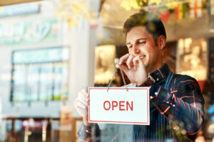A smiling small business owner hangs an 'OPEN' sign on the inside of a glass window, signaling the start of business hours. The image reflects a welcoming atmosphere, symbolizing opportunities like small business grants to help entrepreneurs thrive.
