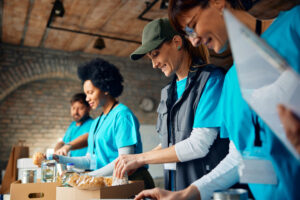 Four volunteers packing food items into cardboard boxes in a rustic indoor setting, symbolizing community support for small businesses through charitable and local engagement efforts.