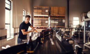 A young man in a black polo shirt and jeans stands next to a conveyor belt filled with amber-colored bottles in a bright, modern manufacturing facility. Representing the spirit of small business, he holds a tablet or clipboard, inspecting the production line. The scene reflects the potential impact of recent house bills, such as H.R. 3174: The Made in America Manufacturing Finance Act and H.R. 2066: The Investing in All of America Act of 2025, which aim to support small manufacturers and boost American production.