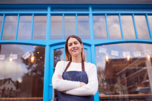 A confident small business owner stands in front of a vibrant blue storefront with large glass windows, wearing a white top and denim apron. The welcoming shop features decorative lights and international flags, symbolizing growth and resilience for small businesses in growing metro areas.