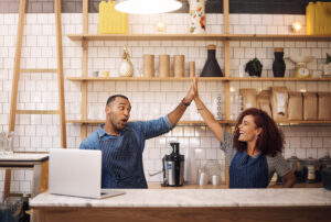 A vibrant scene inside a modern cafe, featuring two baristas in matching aprons celebrating with a high-five over a marble counter. The counter holds a laptop, a blender, and stacked plastic cups, symbolizing teamwork and success. The background showcases neatly arranged shelves with decorative items, plants, and supplies, creating a welcoming atmosphere. This image represents the excitement and opportunities that small business grants can bring to entrepreneurs.