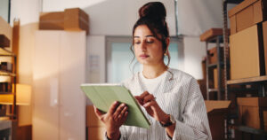A young woman in a storage area holding a tablet, surrounded by shelves of cardboard boxes, representing inventory management and preparedness. The scene highlights the importance of organizing emergency supplies for small businesses.