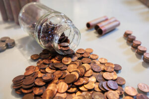 A clear glass jar tipped over on a light surface, spilling numerous copper pennies, with small stacks of coins and rolled penny wrappers in the background. Represents the end of the penny and the impact of penny production cessation for small businesses.