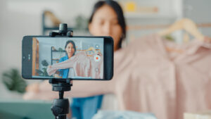 A woman presenting a pink shirt on a hanger while being recorded on a smartphone mounted on a tripod, symbolizing content creation for small business and social media strategies 2026.